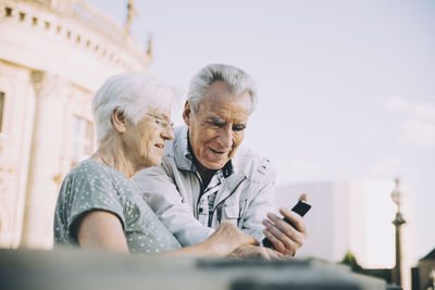 Senior male and female tourists using smart phone while standing in city against sky