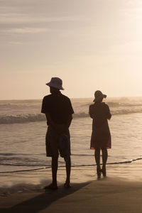 Rear view of people standing at beach against sky during sunset