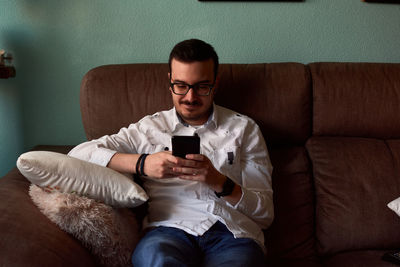 Young man writes in his phone sitting in a couch at home