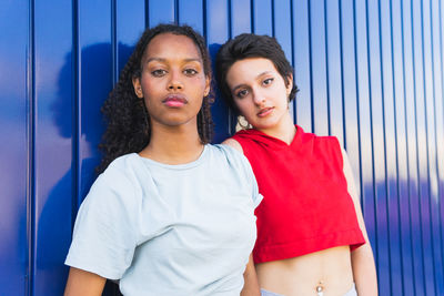 Serious multiracial young female friends in casual clothes looking at camera while standing together against blue background on street of city