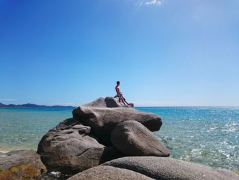 Man sitting on rocks at beach against blue sky