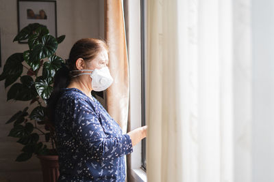 Woman wearing mask looking through window at home