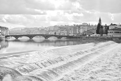 Arch bridge over river amidst buildings against cloudy sky