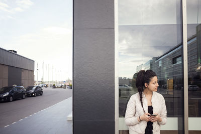 Young woman using smart phone while looking away against window
