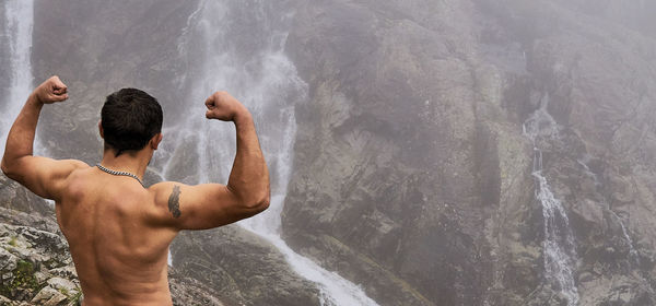 Rear view of shirtless muscular man flexing muscles while looking at waterfall