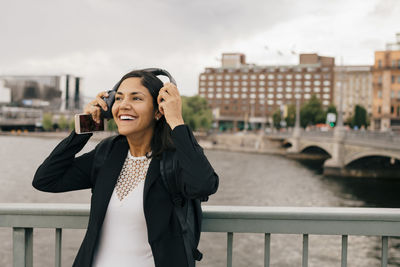 Smiling businesswoman wearing headphones while standing on bridge in city