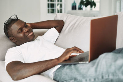 Smiling man using laptop while lying down on sofa at home