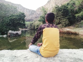 Rear view of man sitting on rock by lake