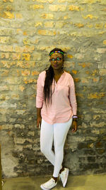 Full length of young woman looking away while standing against old brick wall