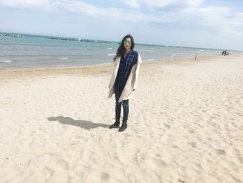 Portrait of young woman wearing sunglasses while standing at beach against sky during sunny day