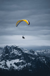 Anonymous person flying on colorful paraglider above snowy mountain range covered with coniferous forest under cloudy gloomy sky person