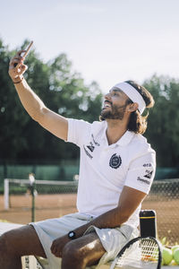 Smiling man taking selfie through smart phone while sitting on bench at tennis court