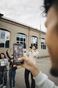 Young man photographing friends through mobile phone on street in city