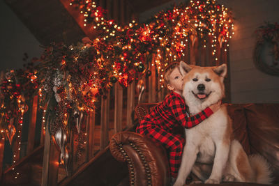 Candid authentic happy little boy in red plaid pajama hugs dog with bow tie at lodge xmas decorated