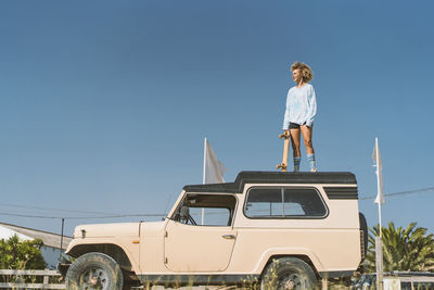 Young woman holding skateboard while standing on old off-road vehicle against blue sky during sunny day
