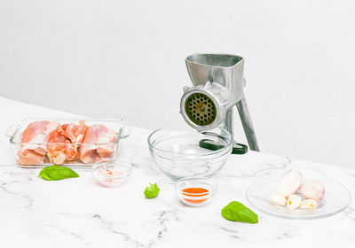 Manual meat grinder with glass bowl,boneless chicken thighs,peeled onions,garlic on a marble table .