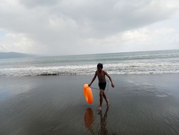 Full length of shirtless boy with inflatable ring walking on shore at beach against cloudy sky