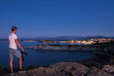 Side view of mid adult man holding camera while standing at beach against clear sky during dusk