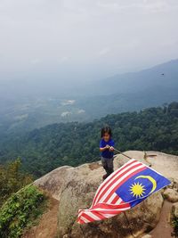 High angle view of boy holding flag while standing on rock against sky