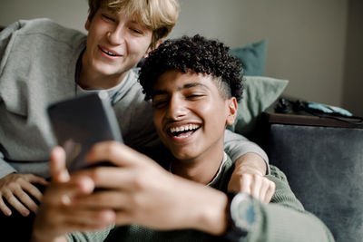 Smiling teenage boy text messaging through smart phone while sitting with friend at home