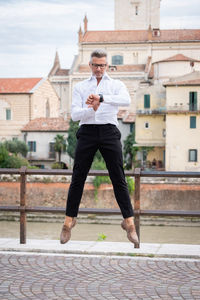 Full length of man jumping against building while checking time in watch