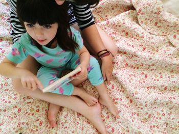 High angle view of cute girl holding book while sitting on mother laps at bed