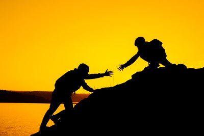 Silhouette girl assisting friends in climbing rock against clear sky during sunset