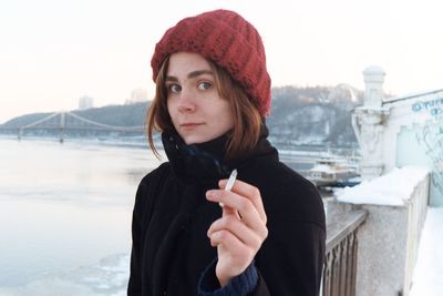 Portrait of young woman with cigarette standing at lake against sky