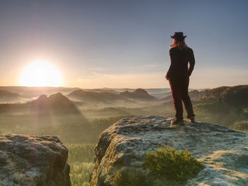 Cheering young woman traveler hiking on rocky mountain promontory above sharp tree tops.