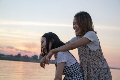 Woman piggybacking friend while standing at beach against sky during sunset