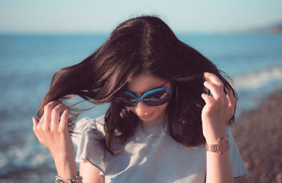 Young woman playing with hair while standing on shore by sea against sky