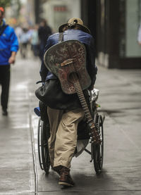 Rear view full length of man with guitar pushing wheelchair on sidewalk