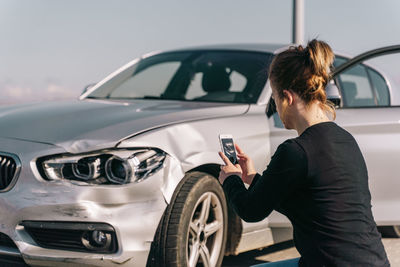 Side view of unrecognizable young female driver in casual clothes taking photo of car damages on smartphone after accident on road on sunny day