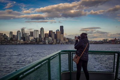 Rear view of woman photographing river against cloudy sky during sunset