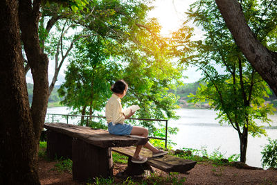 Boy sitting on seat by lake against trees