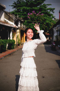 Smiling young woman waving while standing on footpath