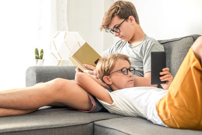 Teenage boy reading book and boy using mobile phone on sofa at home 