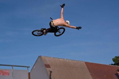 Low angle view of shirtless man performing mid-air stunt with bicycle against blue sky in skateboard park