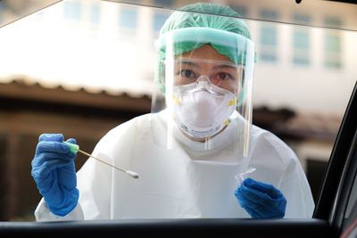 Portrait of doctor wearing protective workwear holding medical equipment seen through car window