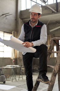 Male engineer holding blueprint looking away while standing on ladder in building