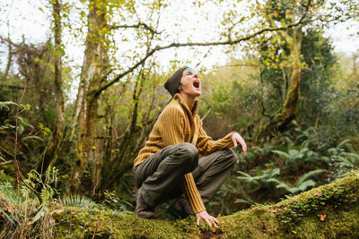 Side view of traveling female pretending being wild animal and roaring with opened mouth while sitting on tree in woods