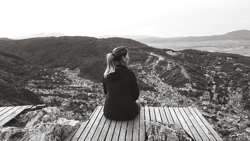 Rear view of mid adult woman looking at mountains while sitting on observation point against clear sky