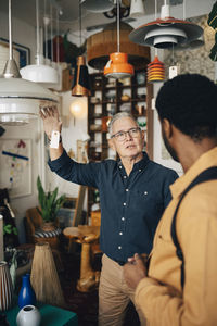 Senior male owner telling price of pendant light to customer standing at antique store