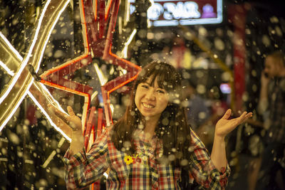 Portrait of smiling woman standing by illuminated decoration during snowfall at night