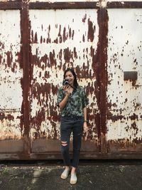 Full length of woman photographing from camera while standing against metallic wall