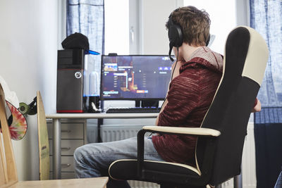 Teenage boy wearing headphones using computer while sitting on chair at home