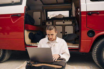 Young man surfing net through laptop while sitting near van during vacation