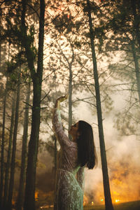 Side view of young woman standing against trees at forest