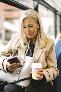 Female professional using smart phone holding disposable cup while sitting in bus