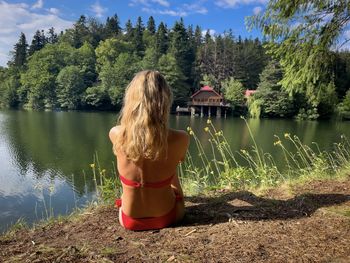 Blonde woman in swimwear sitting on the ground and looking at the lake surrounded by forest 